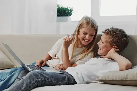 Двое улыбающихся детей отдыхают на диване, пользуясь ноутбуком и наслаждаясь временем, проведенным вместе в помещении