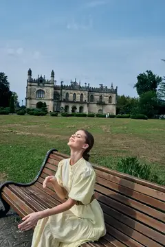 Молодая женщина в светлом платье отдыхает на скамейке в парке у старинного замка