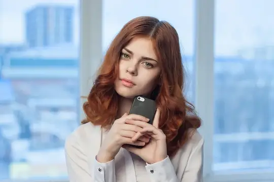 Рыжеволосая девушка держит телефон у окна с видом на город