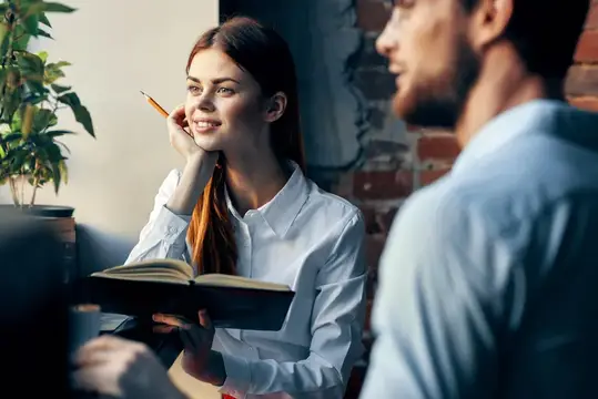 Улыбчивая девушка с книгой и карандашом слушает мужчину в кафе