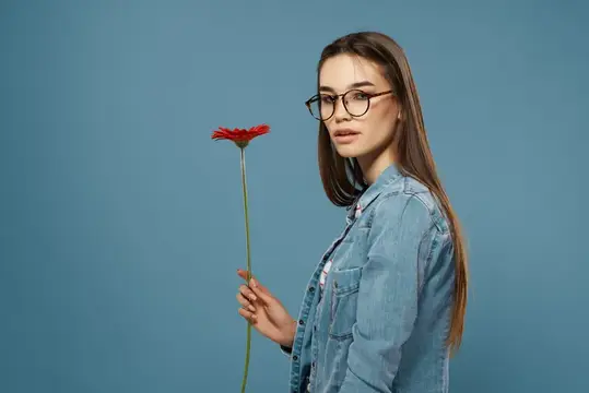 Молодая девушка в очках и джинсовой куртке держит красный цветок на синем фоне