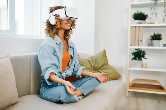 Кудрявая девушка в VR-очках сидит на диване со скрещёнными ногами