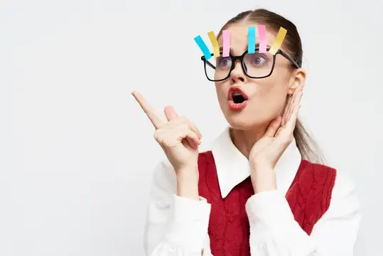 Удивленная женщина в очках, украшенных разноцветными скрепками, восторженно указывает пальцем, выражая всплеск креативности и генерации идей на фоне однотонного белого фона, демонстрируя радость и…