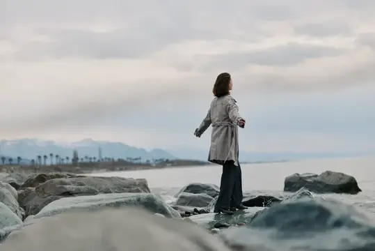 Женщина в плаще стоит на камнях у моря под облачным небом
