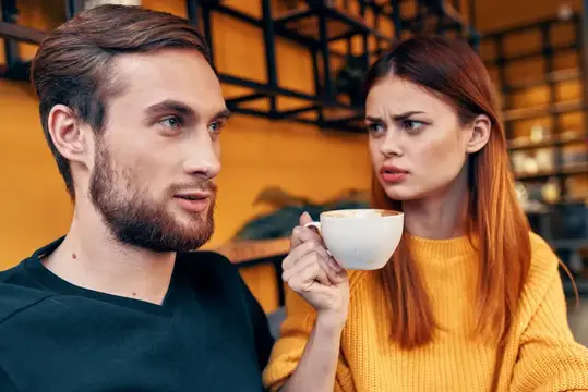 Рыжеволосая женщина с чашкой кофе недоверчиво смотрит на бородатого мужчину в кафе
