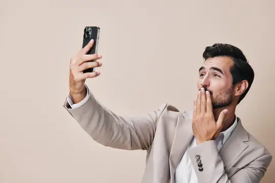 Бородатый мужчина в деловом костюме держит смартфон и улыбается, стоя в студии на сером фоне
