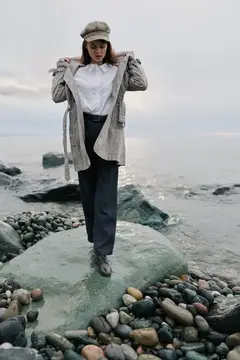Молодая женщина в кепке и плаще стоит на камне у моря
