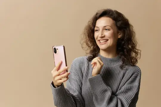 Кудрявая женщина в сером свитере улыбается, глядя в смартфон