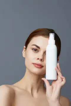 Молодая женщина с белым флаконом косметики у лица на сером фоне