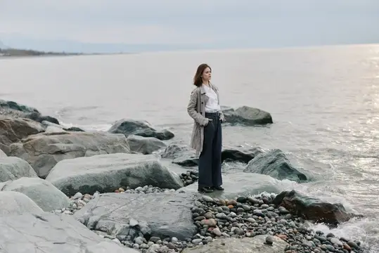 Молодая женщина в плаще стоит на камнях у моря