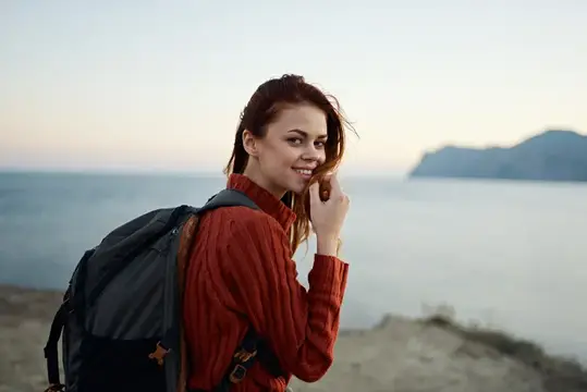Женщина-туристка с рюкзаком и в красном свитере гуляет по пляжу у моря в горах