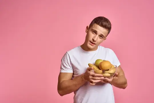 Молодой мужчина с бананами и яблоком в руках на розовом фоне