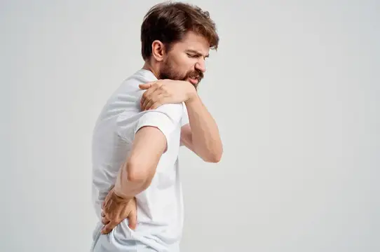 Мужчина в белой футболке массирует плечо и морщится от боли