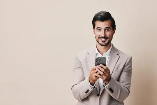 Улыбающийся мужчина в бежевом пиджаке держит смартфон на светлом фоне