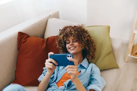 Кудрявая девушка с улыбкой смотрит в смартфон на диване