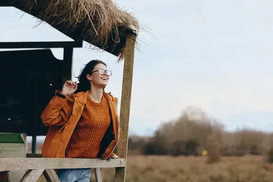 Улыбающаяся девушка в очках и оранжевой куртке у деревянной беседки