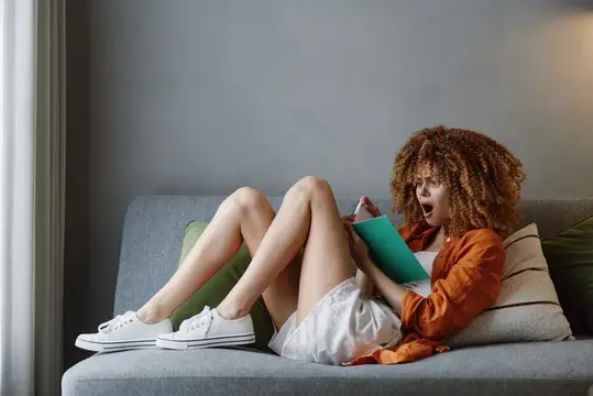 Кудрявая девушка с книгой удивлённо лежит на диване