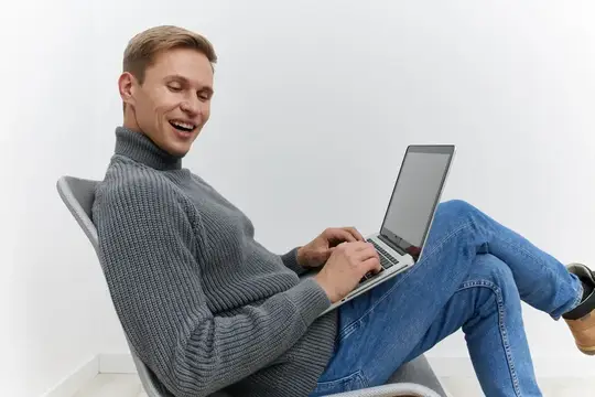 Улыбающийся мужчина в сером свитере работает за ноутбуком в кресле