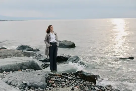 Молодая женщина в плаще стоит на камнях у моря