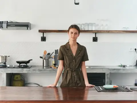 Молодая женщина в платье стоит за деревянным столом на светлой кухне