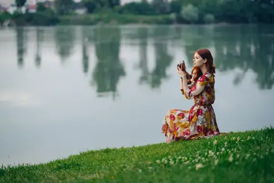 Рыжеволосая женщина в цветастом платье фотографирует на берегу озера