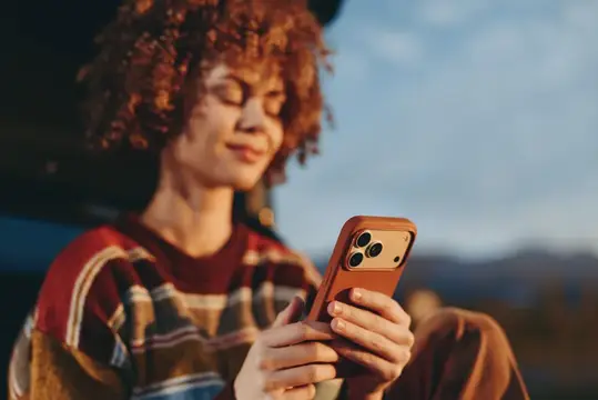 Рыжеволосая девушка с кудрями улыбается, глядя в смартфон на закате