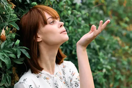 Рыжеволосая девушка с ладонью у лица на фоне зелёных кустов