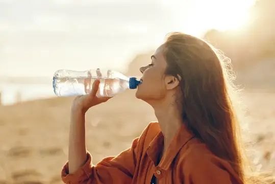 Молодая женщина пьёт воду из бутылки на закатном пляже