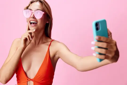 Стильная женщина в купальнике делает селфи на смартфон на фоне яркого розового фона