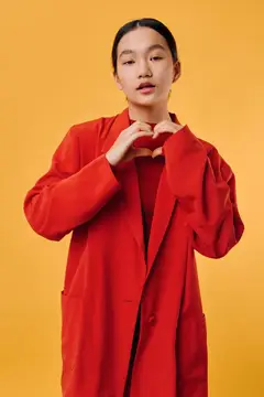 Молодая девушка в красном пальто oversized позирует на однотонном жёлтом фоне, показывая жест «сердце» руками