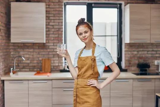 Молодая женщина в фартуке с стаканом воды на кухне