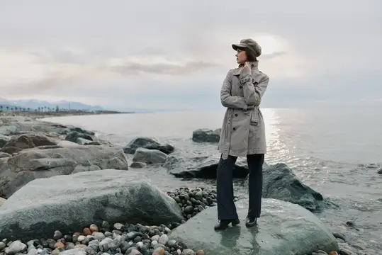 Женщина в плаще и кепке стоит на камнях у моря в пасмурный день