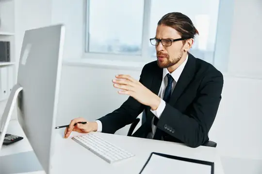Мужчина в очках и костюме жестикулирует перед монитором в офисе