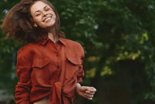 Улыбающаяся женщина на улице в рубашке цвета жженого апельсина запечатлевает жизненный момент при естественном освещении на фоне парка, передавая счастье, энергию, благополучие и повседневную моду в…