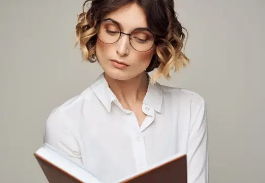 Молодая женщина в очках и белой рубашке читает книгу