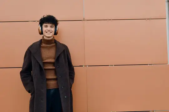 Улыбающийся парень в наушниках и пальто на фоне терракотовой стены