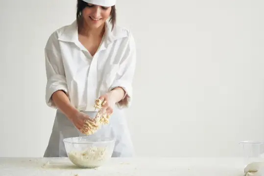 Улыбающаяся женщина в белом фартуке замешивает тесто руками