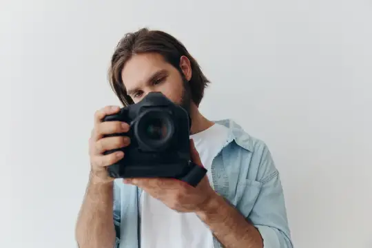 Мужчина-фотограф в стиле хипстер в студии на белом фоне держит профессиональную камеру и настраивает её перед съёмкой