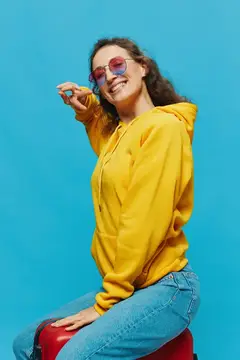 Улыбчивая девушка в жёлтом худи и розовых очках сидит на красном чемодане