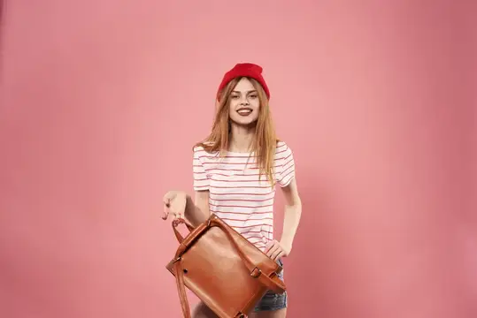 Улыбчивая девушка в красном берете держит собачку в сумке на розовом фоне