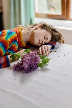 Молодая женщина в радужном свитере лежит на столе с веткой сирени
