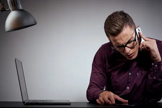 Бизнесмен в очках и рубашке сидит за столом с открытыми ноутбуками, кофе, свободное пространство, мобильный телефон