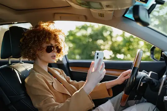 Стильная водительница на сиденье автомобиля сосредоточена на смартфоне и дороге впереди, что передаёт расслабленное настроение, городской пейзаж и современный образ жизни через ретро-плёночную…