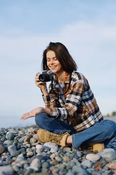 Улыбающаяся девушка с фотокамерой сидит на каменистом берегу
