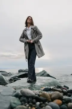 Женщина в плаще стоит на камне у моря в пасмурный день
