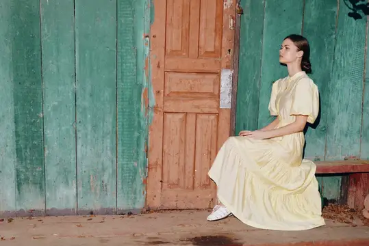 Молодая женщина в бледно-желтом платье сидит у деревянной двери