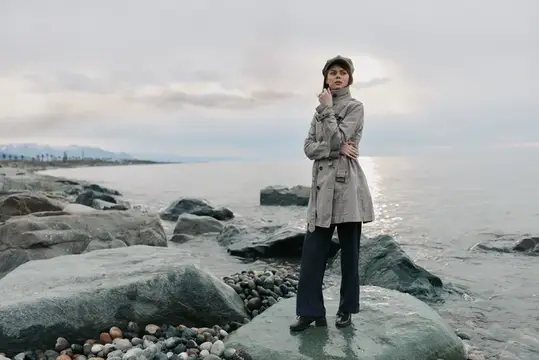 Молодая женщина в плаще и кепке стоит на камнях у моря