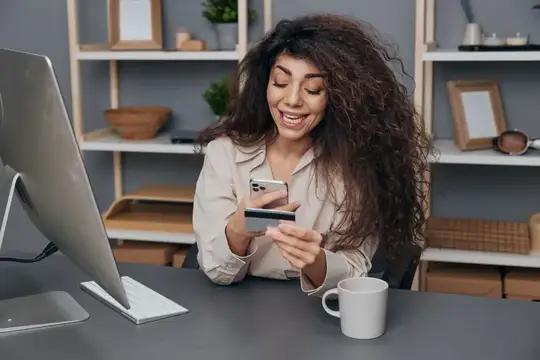 Кудрявая женщина с телефоном и картой улыбается за столом с компьютером