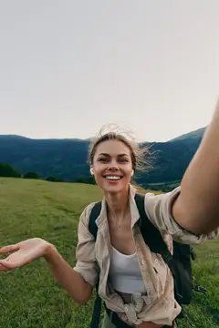 Улыбающаяся девушка с рюкзаком делает селфи на фоне гор