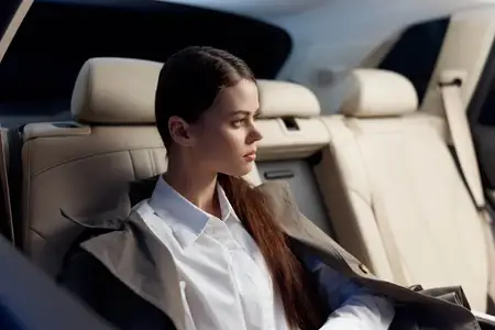Женщина сидит на заднем сиденье автомобиля, положив руки на колени, и отводит взгляд в сторону